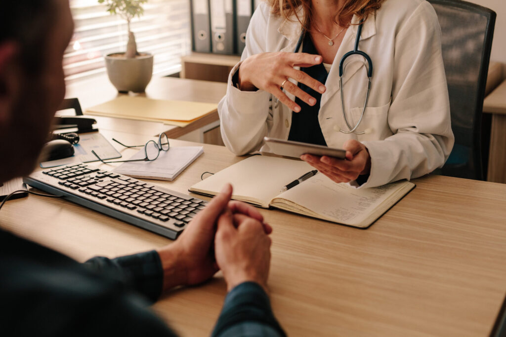 Ärztin mit Stethoskop erklärt einem Patienten am Schreibtisch medizinische Informationen anhand eines Tablets