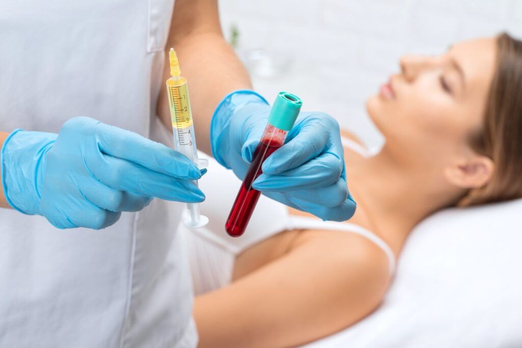 PRP Behandlung: Medizinische Einordnung, Ablauf und realistische Erwartungen Eine medizinische Fachkraft mit Handschuhen hält eine Spritze und ein Röhrchen mit Blut, während eine Frau im Hintergrund auf einer Behandlungsliege liegt.